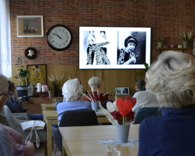Sala. Seniorzy siedzą przy stolikach. Patrzą w stronę telewizora. Na ekranie czarno-białe zdjęcia z aktorka Ireną Kwiatkowską.