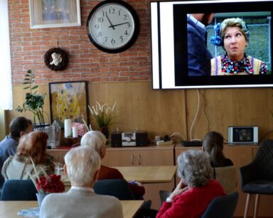 Sala. Seniorzy siedzą przy stolikach. Patrzą w stronę telewizora. Na ekranie aktorka Irena Kwiatkowska.