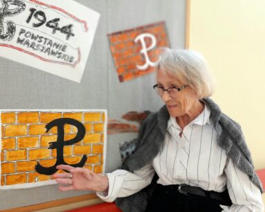 Pomieszczenie. Przy tablicy z plakatami stoi seniorka. Wskazuje ręka plakat z symbolem Polski Walczącej.