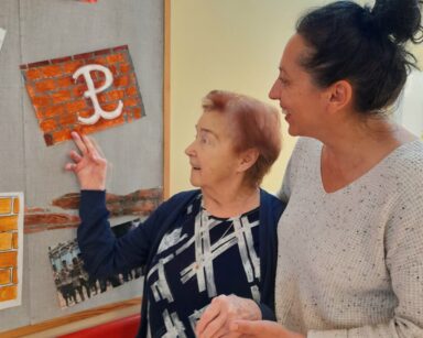 Pomieszczenie. Przy tablicy z plakatami stoi seniorka i terapeutka Magdalena Poraj-Górska. Seniorka wskazuje ręka plakat z symbolem Polski Walczącej.