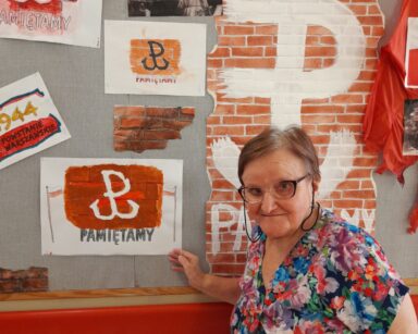Seniorka pozuje do zdjęcia przed tablicą. Na tablicy napis Powstanie i plakaty z symbolami Polski Walczącej.