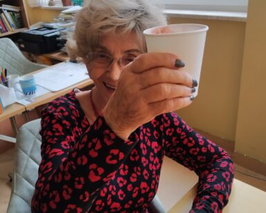 Sala. Seniorka pozuje do zdjęcia. Siedzi przy stole. Śmieje się. Unosi kubek w geście toastu.