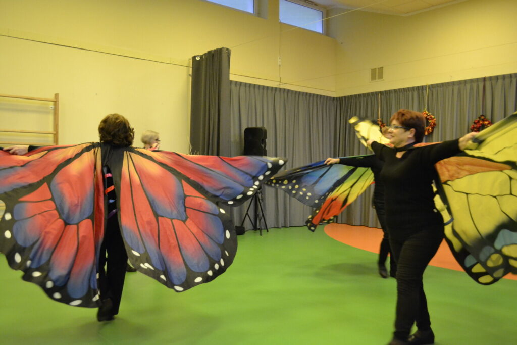 Sala. Trzy kobiety ubrane w skrzydła motyla tańczą.