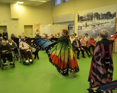 Sala. Grupa seniorów ogląda występ grupy tanecznej. Dwie kobiety mają na sobie strój motyla.