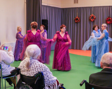 Sala. Sześć kobiet tańczy. Ubrane są w kolorowe długie suknie. Grupa seniorów ogląda występ.