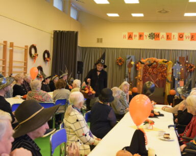 Sala. Grupa seniorów w przebraniu Halloween siedzi przy stole. Kobieta w czerni trzyma w ręku kosz.