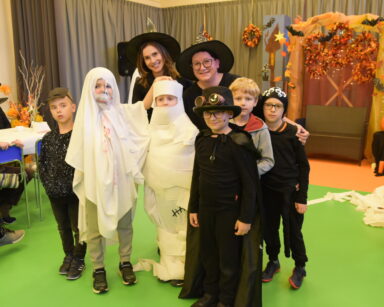 Sala. Grupa pierwszoklasistów pozuje do zdjęcia. Maja na sobie ubrania Halloween. W tle dwie kobiety w czerni.
