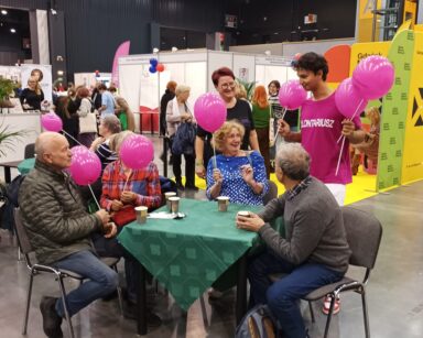 Sala. Czterech seniorów siedzi przy stole i trzyma różowe balony. Obok stoją wolontariusze z Sopockiego Centrum Seniora.