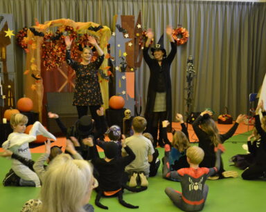 Zbliżenie. Scena. Dwie kobiety unoszą ręce do góry. Przed sceną siedzi grupa dzieci. W tle ozdoby Halloween.