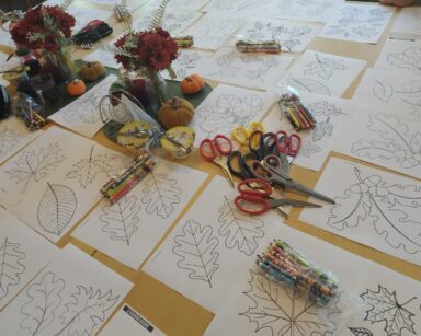 Zbliżenie. Stół. Na stole leżą kartki z rysunkami liści. Obok kredki i nożyczki.