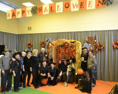 Sala. Grupa osób przebrana w stroje Halloween pozuje do zdjęcia. W tle napis Happy Halloween.
