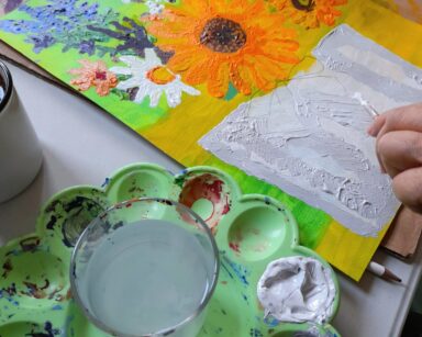 Zbliżenie. Stół. Na stole stoi kubek z wodą i farbki. Obok Kobieta maluje obraz.