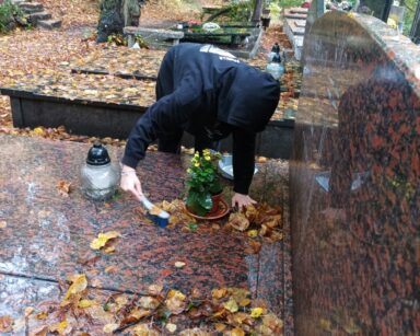 Cmentarz. Wolontariusz pochylony nad grobem. Sprząta z grobu liście.