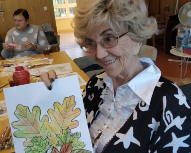 Zbliżenie. Seniorka pozuje do zdjęcia. W ręku trzyma wykonaną przez siebie jesienna kompozycję.