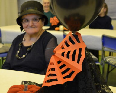 Zbliżenie. Na stole stoi stroik. Czarny balon, koronka i pomarańczowe dynie. W tle siedzi seniorka ubrana na czarno.