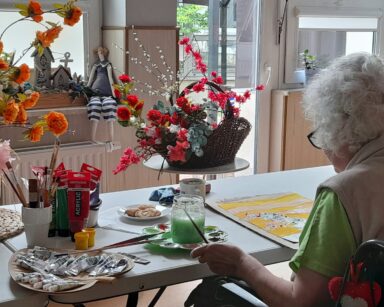 Pokój. Przy oknie stoi kosz z kwiatami. Przy stole siedzi kobieta i maluje obraz.