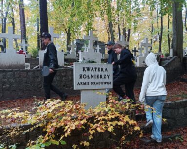 Cmentarz. Czterech wolontariuszy przechodzi koło tablicy. Na niej napis Kwater żołnierzy Armii Krajowej.
