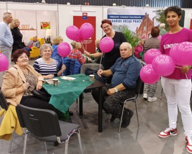 Sala. Trzech seniorów siedzi przy stole. Obok Pani Edyta i Pan Krzysztof z różowymi balonami pozują do zdjęcia.