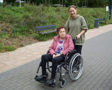 Piękny dzień. Wolontariuszka spaceruje z seniorką. Seniorka porusza się na wózku.