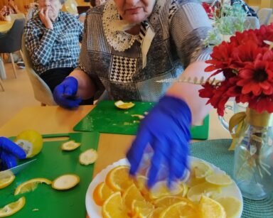 Zbliżenie. Seniorka siedzi przy stole i obiera pomarańczę.