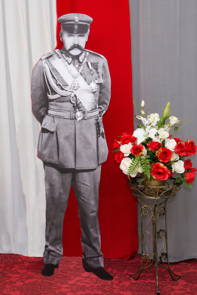 Zbliżenie. Zdjęcie. Postać Marszałka Józefa Piłsudskiego na tle biało czerwonej flagi Polski.