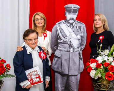 Zbliżenie. Trzy kobiety pozują do zdjęcie. Obok postać Marszałka Piłsudskiego. W tle flaga Polski.
