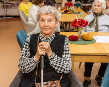 Zbliżenie. Kobieta siedzi na krześle. W ręku trzyma laskę. W tle seniorzy siedzą przy stole.