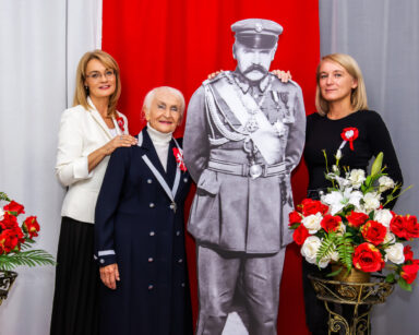 Zbliżenie. Trzy kobiety pozują do zdjęcia z postacią Piłsudskiego. W tle flaga Polski. Obok bukiety kwiatów.