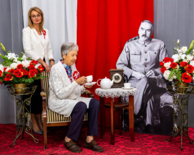 Zbliżenie. Seniorka siedzi na fotelu. Obok siedzi Piłsudski. Za kobietą stoi Dyrektor DPS. W tle flaga Polski.