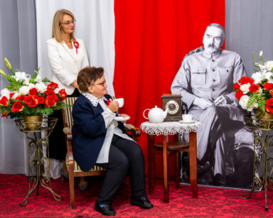 Zbliżenie. Kobieta trzyma w ręku filiżankę. Obok na fotelu siedzi postać Marszałka. Za kobieta stoi Dyrektor DPS.