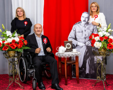 Zbliżenie. Senior siedzi na wózku. Obok na fotelu J. Piłsudski. Za mężczyznami stają dwie kobiety. W tle flaga Polski.