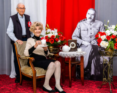 Zbliżenie. Para seniorów pozuje do zdjęcia z Piłsudskim. Kobieta siedzi na fotelu z filiżanką w dłoni za nią stoi senior.
