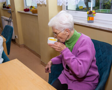 Zbliżenie. Seniorka w fioletowym swetrze siedzi w fotelu. W ręku ma kubek z zupą. Kubek trzyma przy ustach.