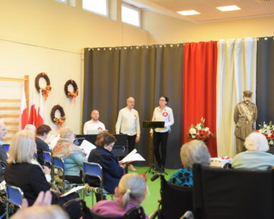 Sala. Grupa seniorów siedzi. Na scenie kierownik DDP. Obok kobieta i mężczyzna stoją przy mikrofonie. W tle flaga Polski.