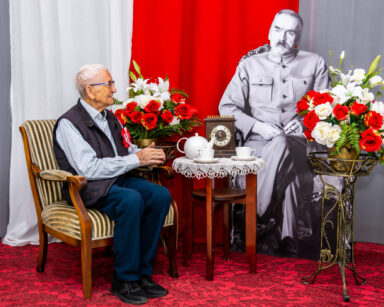 Zbliżenie. Senior siedzi na fotelu. Obok stoi stolik przy którym siedzi Marszałek Piłsudski. W tle flaga Polski.