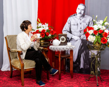 Zbliżenie. Kobieta z filiżanką w dłoni siedzi na fotelu. Obok przy stoliku siedzi Marszałek Piłsudski.