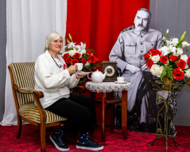 Zbliżenie. Kobieta Z filiżanką siedzi na fotelu. Obok przy stoliku siedzi J. Piłsudski. W tle flaga Polski. Obok kwiaty.
