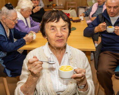 Zbliżenie. Seniorka je zupę z dyni. Jest uśmiechnięta. W tle grupa ludzi siedzi przy stole i jedzą zupę z dyni.
