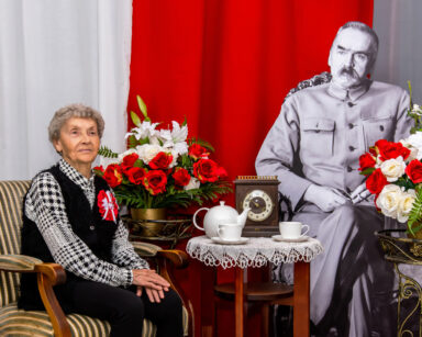 Zbliżenie. Uśmiechnięta kobieta siedzi na fotelu. Obok przy stoliku siedzi Marszałek Piłsudski. W tle flaga Polski.