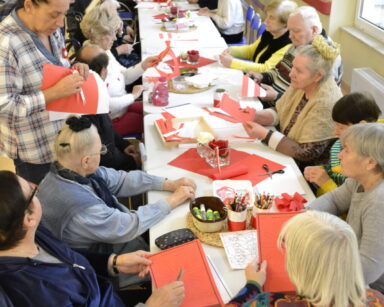 Korytarz. Długi stół. Przy stole siedzą seniorzy. Obok stoją opiekunowie. Na stole leżą czerwone i białe kartki, kredki.
