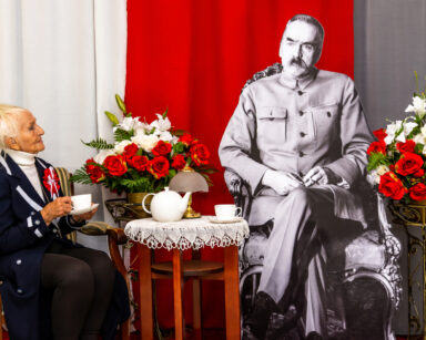 Zbliżenie. Kobieta siedzi z filiżanką przy stoliku, obok siedzi Piłsudski. Na stoliku imbryk. Obok kwiaty biało czerwone.