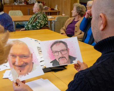 Zbliżenie. Mężczyzna siedzi przy stole. W dłoniach trzyma dwa portrety mężczyzn. Do twarz dorysowane są wąsy.