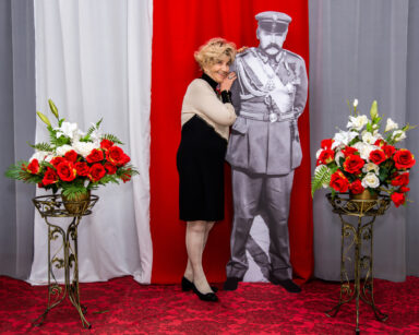 Zbliżenie. Zdjęcie. Uśmiechnięta kobieta opiera się o postać Marszałka Piłsudskiego. Obok kwiaty. W tle flaga Polski.