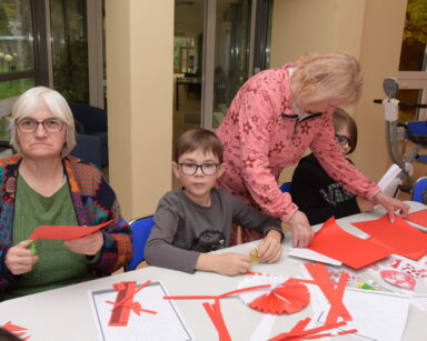 Zbliżenie. Seniorka z dziećmi siedzi przy stole. Druga seniorka pochylona nad stołem trzyma w rękach czerwone kartki.