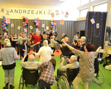 Sala. Grupa seniorów bawi się na sali. Na scenie tańczą młodzi ludzie. W tle napis Andrzejki.