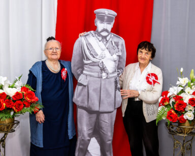 Zbliżenie. Dwie kobiety pozują do zdjęcia z postacią Marszałka Piłsudskiego. Obok kwiaty. W tle flaga Polski.