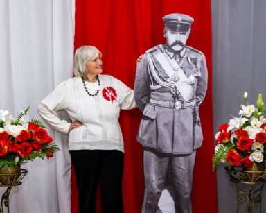 Zbliżenie. Kobieta z ręka na ramieniu Marszałka Piłsudskiego pozuje do zdjęcia. Obok kwiaty. W tle flaga Polski.