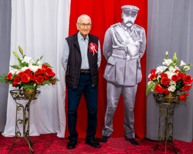Zbliżenie. Senior pozuje do zdjęcia z Marszałkiem Piłsudskim. Panowie maja ręce splecione z tyłu. W tle flaga Polski.