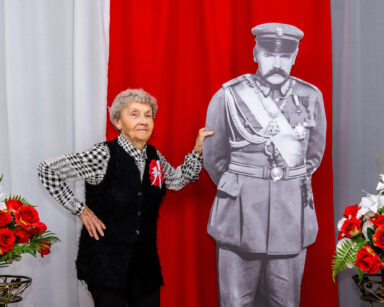 Zbliżenie. Kobieta opiera się ręka o postać Piłsudskiego. Drugą trzyma na boku. Pozuje do zdjęcia. W tle flaga Polski.