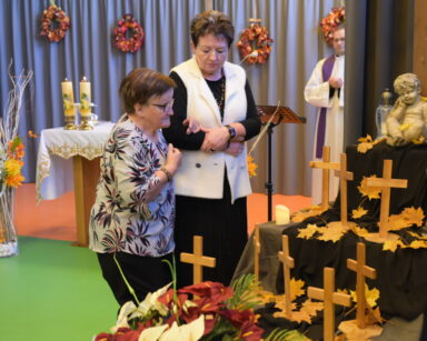 Zbliżenie. Dwie kobiety stoją przy konstrukcji z krzyżami. Obok stoją znicza i figurka anioła. W tle stoi ksiądz.